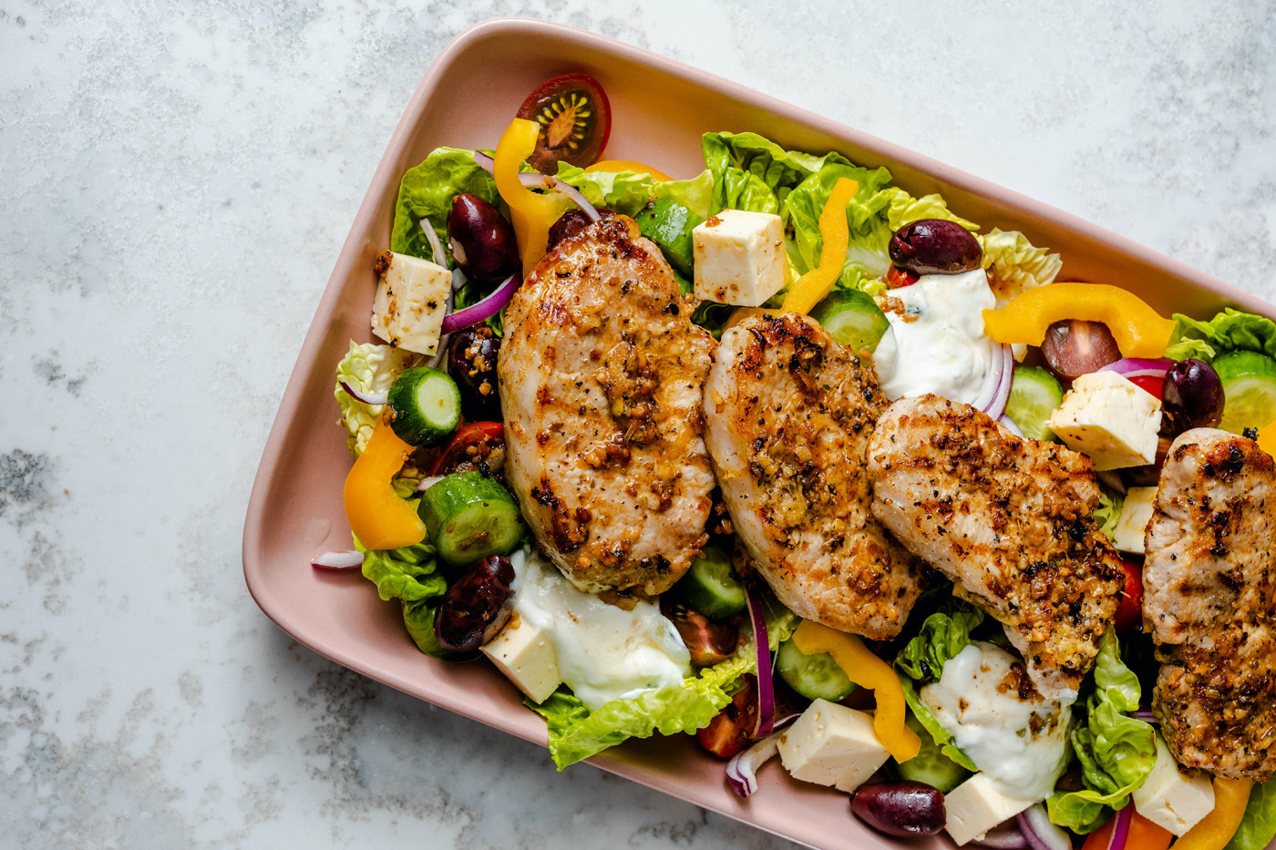 Juicy lean pork steaks on top of a classic Greek salad with baby gem lettuce leaves, Kalamata olives, yellow capsicum, cherry tomatoes, red onion, Greek feta and Tzatziki. Served on a light pink rectangular serving plate, on a marble bench top